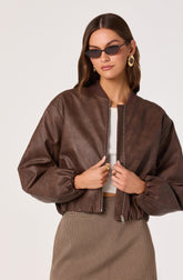 A woman is wearing a brown leather bomber jacket with a front zipper, a white ribbed crop top underneath, and high-waisted checkered pants. She has on large, round earrings, dark sunglasses, and a ring on each hand.